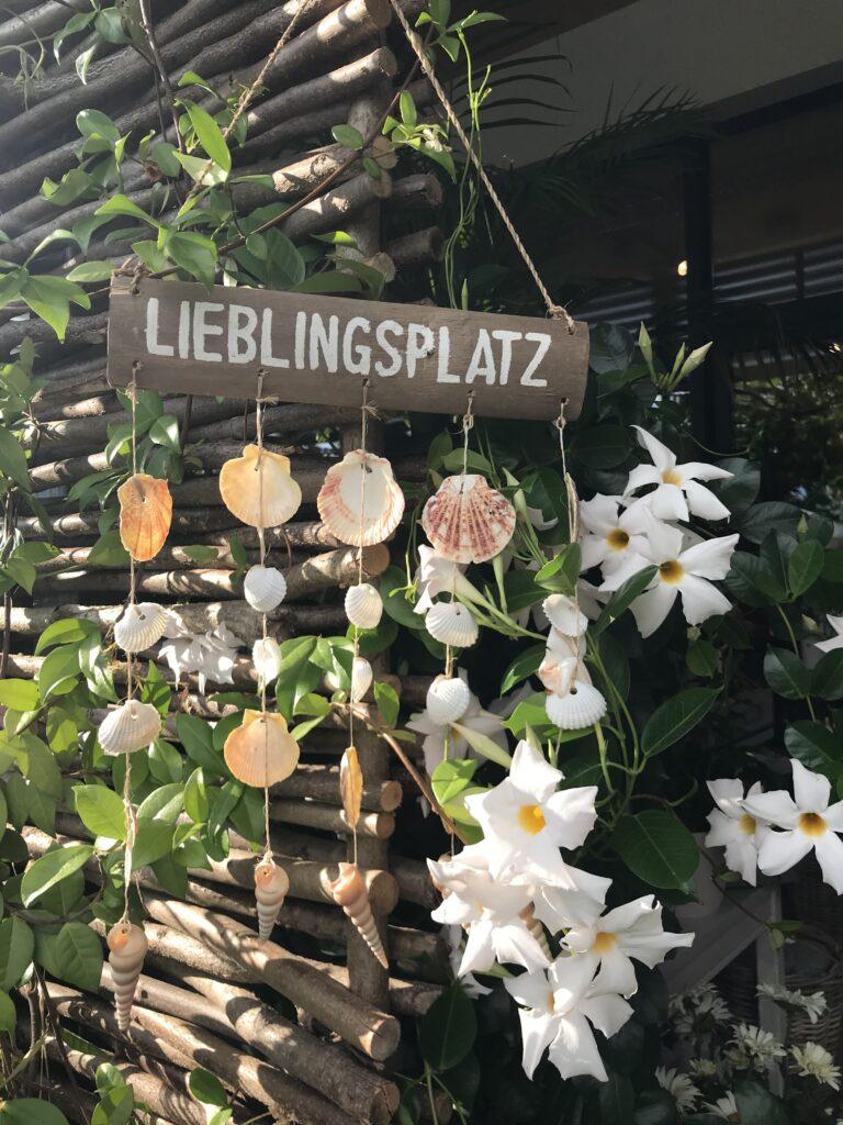 Holzschild mit der Aufschrift 'Lieblingsplatz' und Muscheln, umgeben von Blumen und Pflanzen